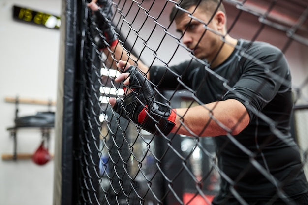 Il maschio bruna vestito di nero e guanti pratica kickboxing, concetto di sport mma, sta in piedi esausto e stanco dopo aver combattuto. Ritratto di vista laterale. Riposati, guarda attraverso la gabbia. concentrati sulle mani nei guanti