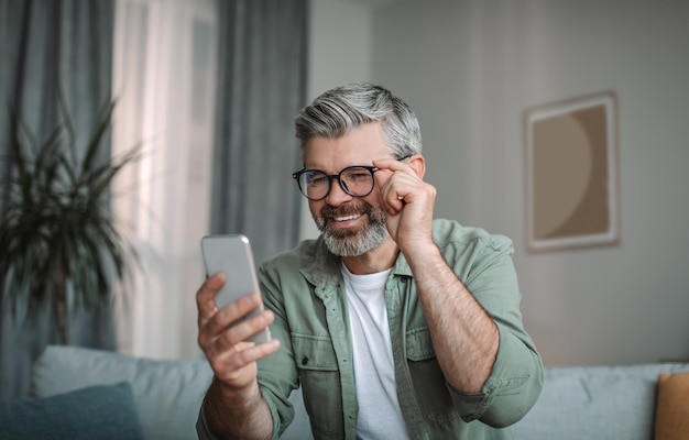 Il maschio caucasico anziano felice con la barba con gli occhiali guarda lo smartphone ha ricevuto buone notizie nello spazio della copia interna della stanza