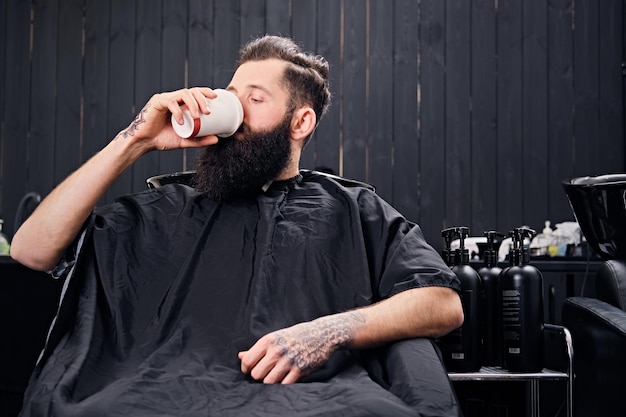 Il maschio hipster barbuto a tutto gas beve il caffè prima dell'acconciatura nel salone di un parrucchiere.