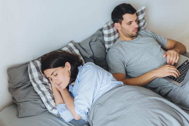 Il maschio impegnato si trova a letto lavora sul computer portatile ha un'espressione seria mentre sua moglie ha un sonno sano stare insieme in camera da letto Due persone a letto Concetto di casa di famiglia delle persone