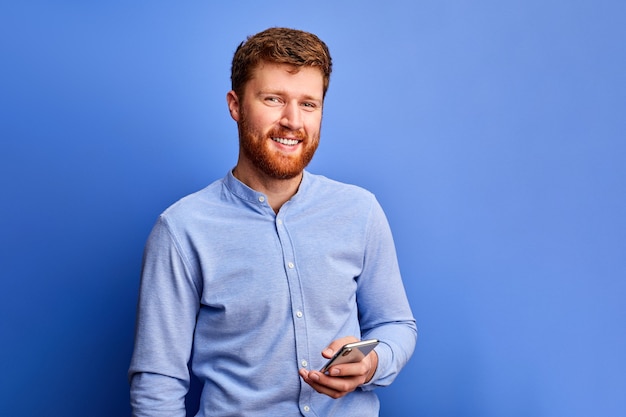 Il maschio ottimista sta chattando con un amico sul cellulare, digitando un messaggio, indossa una camicia formale blu, sorridendo