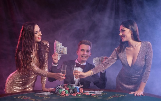 Il maschio sta giocando a poker al casinò seduto al tavolo con pile di fiches carte denaro Celebrando la vittoria con due signore sorridenti Sfondo fumo nero retroilluminazioni colorate Gioco d'azzardo alcol Primo piano