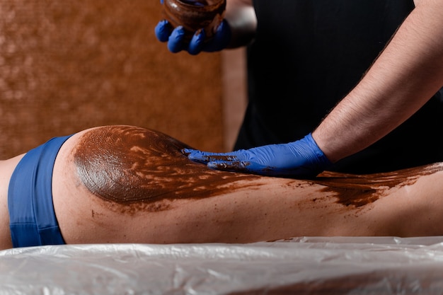 Il massaggiatore spalma di cioccolato le gambe e le cosce della ragazza nel centro termale. Impacco al cioccolato con procedura di bellezza.