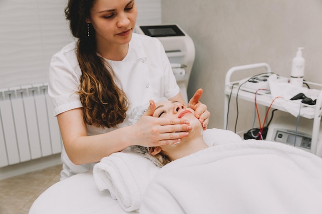 Il massaggio facciale viene preparato nella spa Mani del medico Trattamento per una pelle sana Testa di giovane donna Detergente ringiovanimento Maschera viso dermatologica Terapia disintossicante