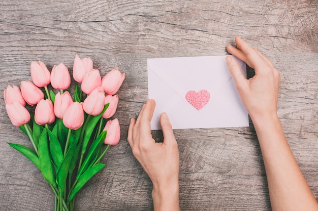 il mazzo dei tulipani rosa e le mani delle donne tengono una busta in bianco con un cuore, su un fondo di legno.