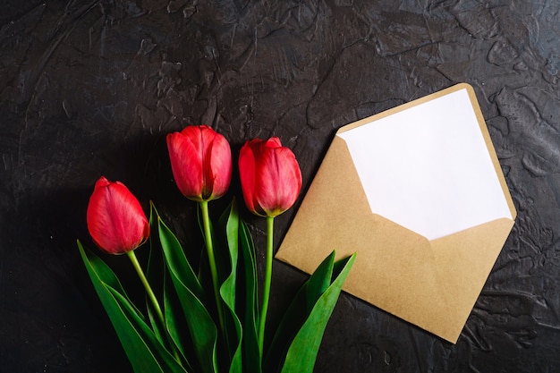 Il mazzo rosso di tulipano fiorisce con la carta della busta su fondo nero strutturato, spazio della copia di vista superiore