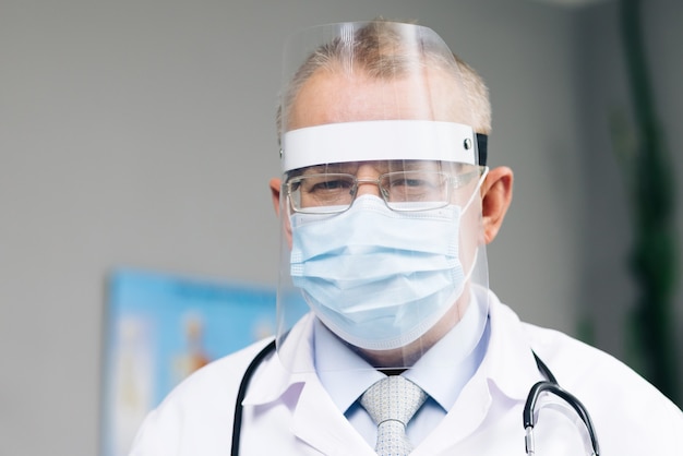 Il medico caucasico con gli occhiali indossa una maschera protettiva trasparente per il viso e una tuta in una stanza d'ospedale