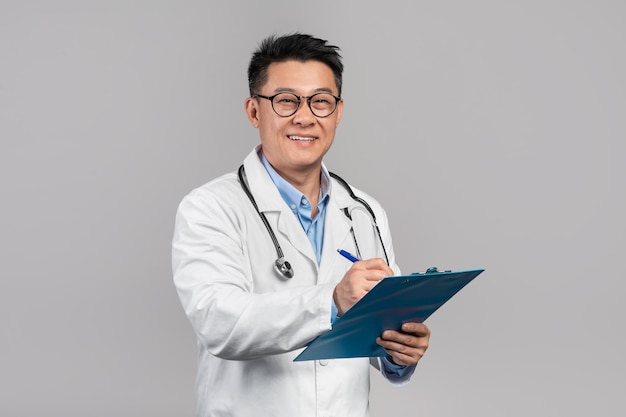 Il medico coreano maturo allegro dell'uomo in vetri del camice bianco con lo stetoscopio scrive al tablet