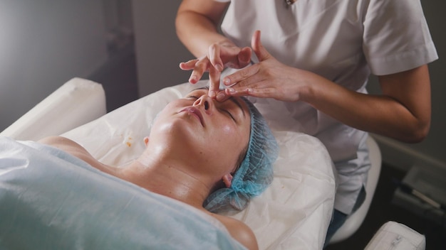 Il medico della donna esegue la procedura cosmetica - massaggio facciale della maschera alla cura della pelle del salone della stazione termale, primo piano