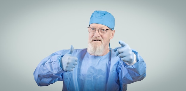 Il medico delle malattie infettive con una barba grigia in una tuta protettiva blu e guanti di gomma mostra un pollice in su