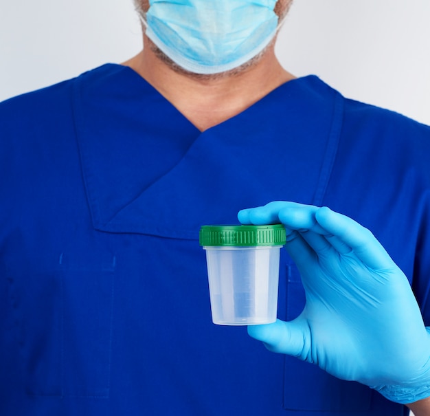 Il medico in uniforme blu e guanti in lattice tiene in mano un contenitore di plastica vuoto per prelevare campioni di urina