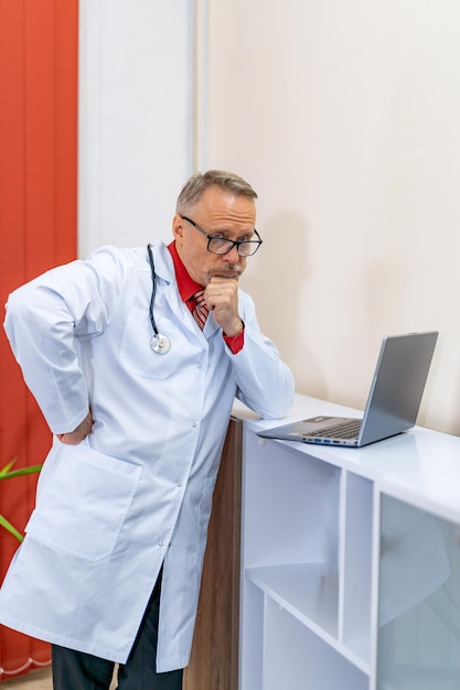 Il medico senior premuroso risolve i problemi sanitari sul laptop Medico professionista in scrub in una clinica moderna