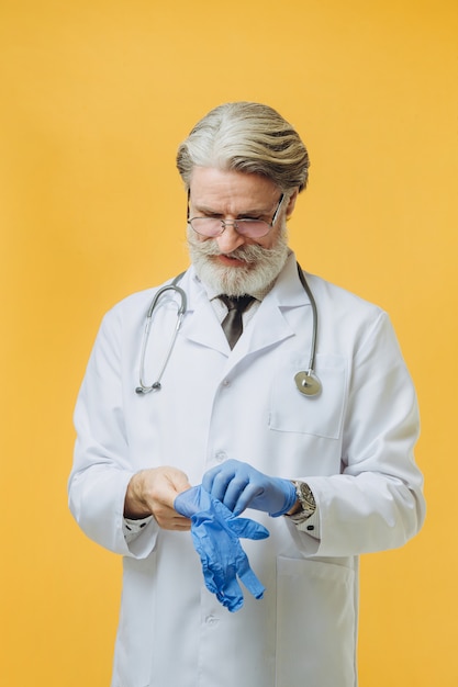 Il medico senor indossa camice bianco e stetoscopio, isolato sul muro giallo, indossa guanti blu