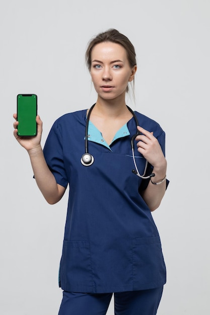 Il medico tiene in mano un telefono cellulare con un chromakey Una giovane donna in tuta da chirurgo blu su sfondo bianco Isolato