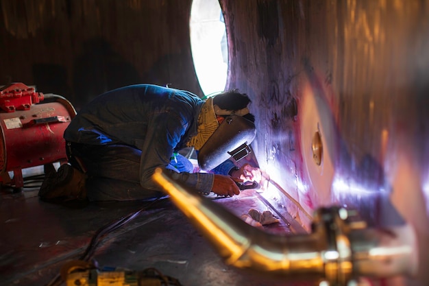 Il metallo riparato maschio del lavoratore dell'argon dell'arco di saldatura sta saldando l'olio inossidabile del serbatoio della costruzione industriale delle scintille all'interno di spazi ristretti.