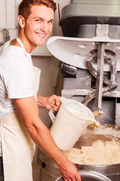 Il miglior impasto per la nostra pasticceria. Fiducioso giovane in grembiule premendo il pulsante sulla macchina impastatrice e sorridente