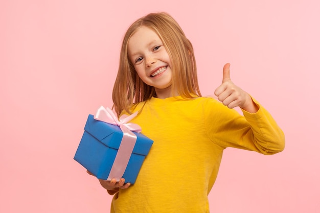 Il miglior regalo. Ritratto di adorabile bambina sorridente che tiene scatola avvolta e mostra i pollici in su, come gesto, bambino soddisfatto del regalo di compleanno. studio indoor girato isolato su sfondo rosa