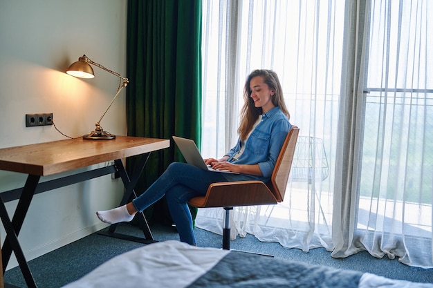 Il millenario sorridenti freelancer ragazza caucasica sedersi alla sedia in camera d'albergo e lavorare a distanza online utilizzando laptop