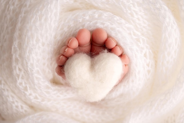 Il minuscolo piede di un neonato. Morbidi piedi di un neonato in una coperta di lana bianca. Primo piano delle dita dei piedi, dei talloni e dei piedi di un neonato. Cuore bianco lavorato a maglia nelle gambe di un bambino. Macrofotografia in studio.