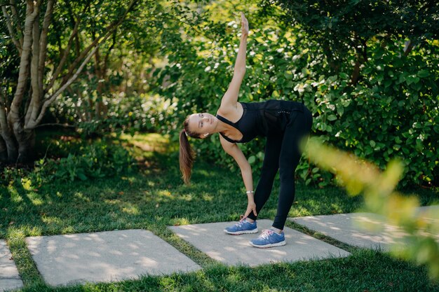 Il modello di fitness attivo si esercita all'aperto si appoggia ai piedi alza il braccio prepara i muscoli prima che l'allenamento cardio si sfida vestita con abbigliamento attivo ha pose di coda di cavallo nel parco verde durante la giornata di sole