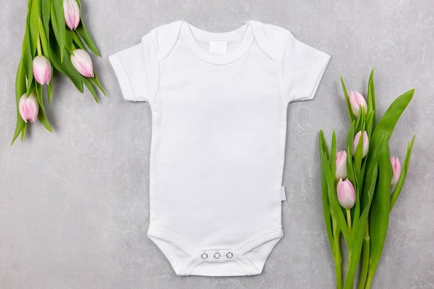 Il modello di mockup di body per neonato bianco giaceva con fiori di tulipani rosa sulla superficie del cemento grigio