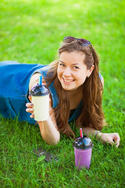 Il modello femminile attraente si trova sul campo in erba e beve il frullato fresco. Giovane donna felice con gustoso milkshake all'aperto.