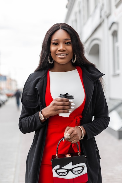 Il modello positivo alla moda della giovane donna africana in vestito rosso alla moda in giacca con la tazza di caffè cammina vicino all'edificio d'epoca in città. La ragazza nera allegra alla moda con un sorriso carino si diverte a bere e camminare.