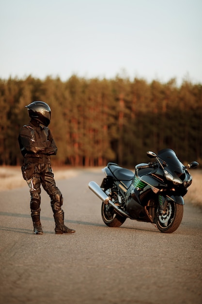 Il motociclista in un casco e una giacca di pelle si trova sulla strada al tramonto con uno sfondo bellissimo bosco e guarda una moto sportiva