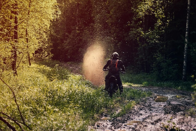 Il motociclista su una moto sportiva di enduro cavalca su una strada fangosa con pozzanghere con schizzi nella foresta in una gara fuoristrada