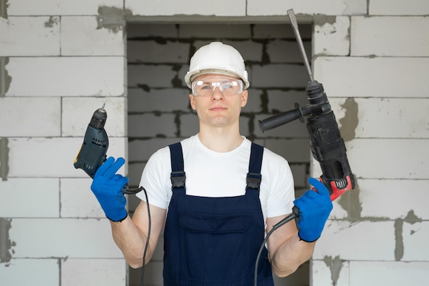 Il muratore professionista con un elmetto bianco tiene in mano un trapano e un cacciavite.