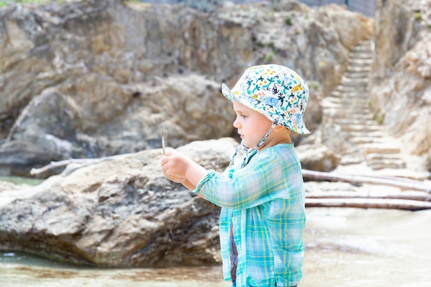 Il neonato del bambino che indossa vestiti di protezione solare e panama sta giocando su una spiaggia rocciosa durante le vacanze estive. Vacanze con il concetto di bambini, copia dello spazio.