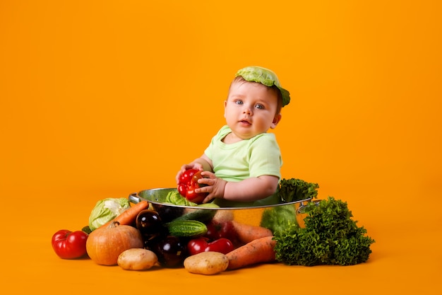 Il neonato in una tuta verde si siede in una ciotola del metallo con le verdure. concetto di prodotto agricolo