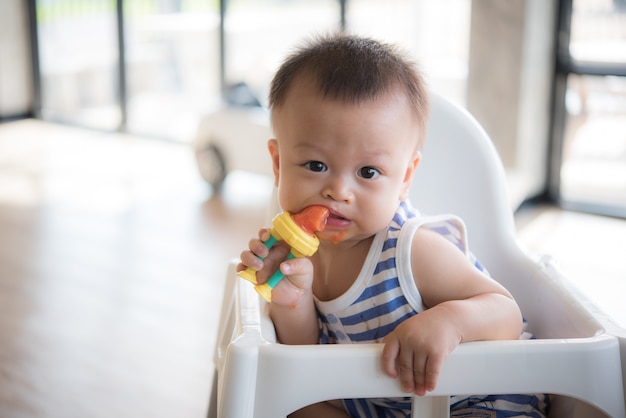 Il neonato sveglio asiatico sta mangiando la frutta dall&#39;accessorio.