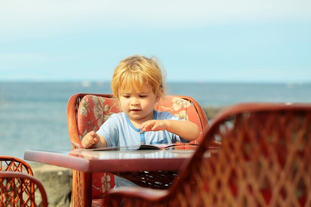 Il neonato sveglio con i capelli biondi in maglietta blu legge la scheda del menu seduto al tavolo nel caffè all'aperto in riva al mare nella soleggiata giornata estiva su sfondo naturale sfocato