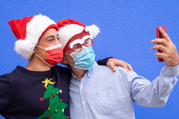 Il nonno con il nipote adolescente sorride usando lo smartphone per fare un selfie, indossando una maschera chirurgica a causa del coronavirus e dei cappelli di babbo natale. Concetto di famiglia, amicizia e divertimento