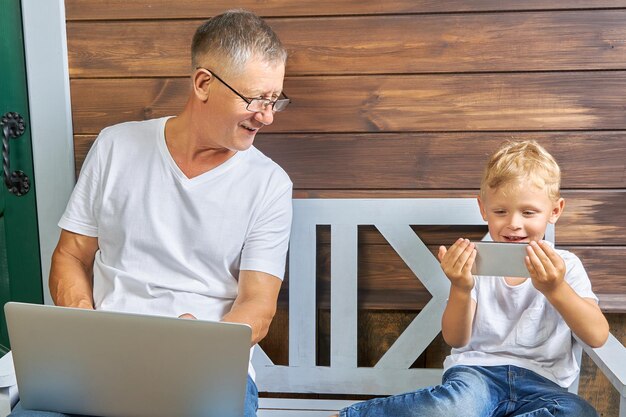 Il nonno con un laptop seduto su una panchina guarda cosa sta facendo suo nipote con uno smartphone vicino a una casa di legno Concetto di utilizzo di gadget moderni di diverse generazioni