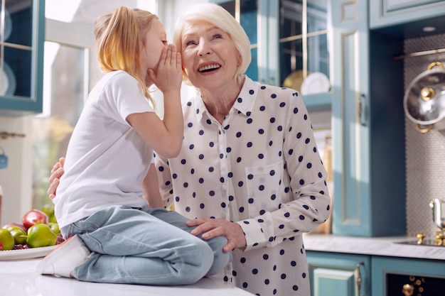 Il nostro segreto. Adorabile bambina seduta sul bancone della cucina e sussurrando all'orecchio della nonna mentre la donna ascolta con attenzione e sorride
