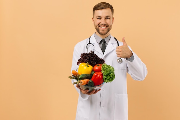Il nutrizionista medico maschio felice con lo stetoscopio mostra le verdure fresche sul concetto di piano di dieta di fondo beige