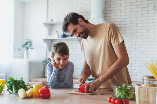 Il padre allegro e il figlio piccolo si aiutano a vicenda mentre preparano un'insalata seduti a un tavolo a casa in cucina