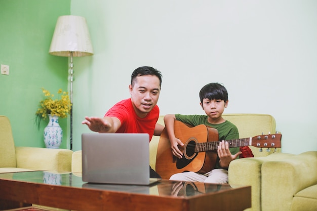 Il padre asiatico mostra come suonare la chitarra a suo figlio con il laptop