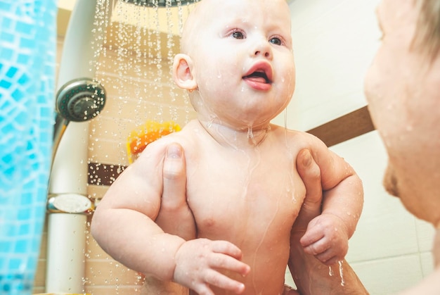 Il padre attento tiene il bambino piccolo sotto l'acqua corrente della doccia nel primo piano del bagno leggero a casa