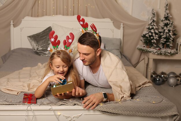 il padre è sdraiato su un letto con una figlia piccola in un bordo di corna di cervo che guarda una scatola regalo di Natale