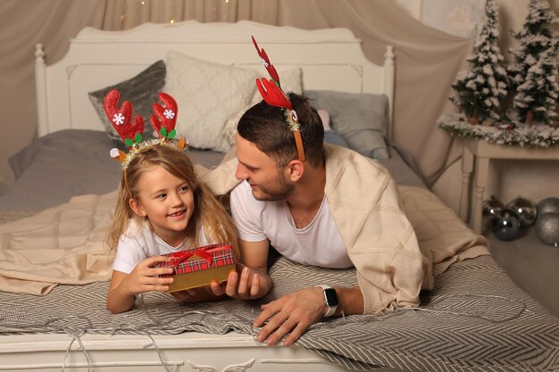 il padre è sdraiato su un letto con una figlia piccola in un bordo di corna di cervo che guarda una scatola regalo di Natale