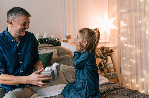 il padre fa un regalo di Natale alla sua piccola figlia seduta sul letto vicino all'albero di Natale