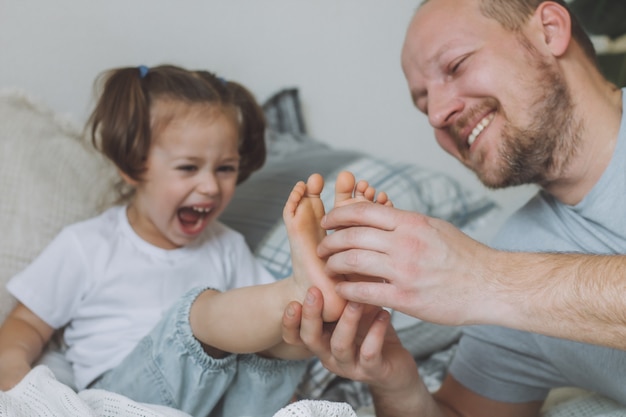 Il padre gioca con la piccola figlia 2-4 sul letto. Papà solletica i piedi dei bambini. Famiglia, divertimento