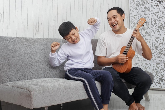 Il padre in maglietta bianca sta suonando l'ukulele mentre canta con suo figlio in maglione sta ballando