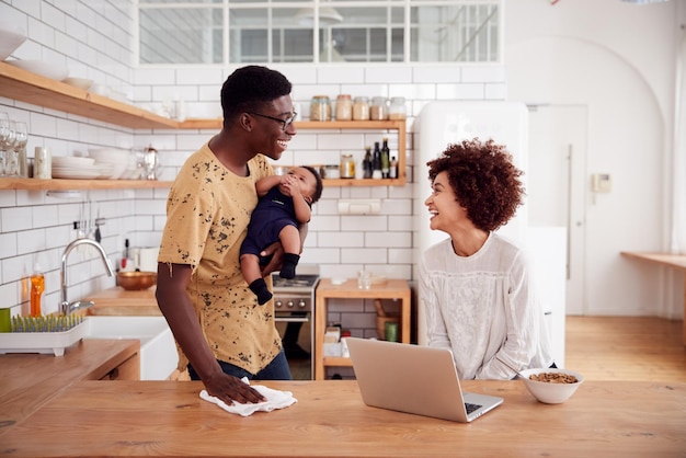 Il padre multitasking tiene il bambino e pulisce la superficie mentre la madre usa il laptop e fa colazione