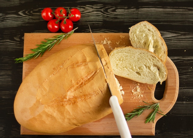 Il pane bianco è affettato con un coltello su una tavola di legno con pomodorini e rosmarino
