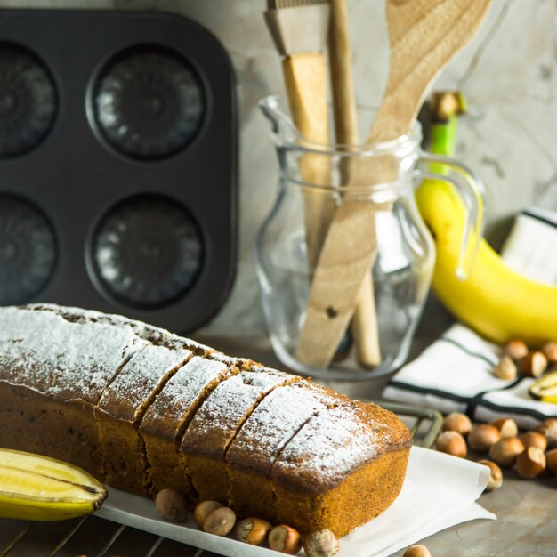 Il pane di banana affettato è sdraiato su una tavola tra utensili da cucina, banane e nocciole