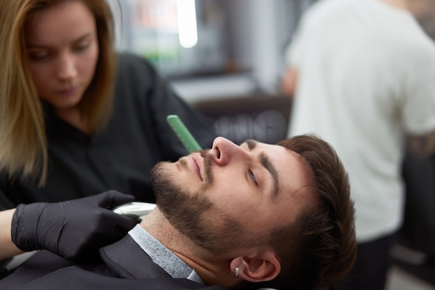 Il parrucchiere della giovane bella donna caucasica taglia l'uomo bello della barba al moderno negozio di barbiere